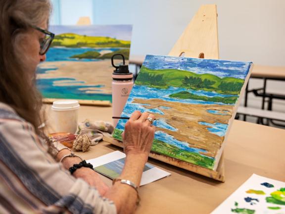 Adult student paints landscape on table top easel with reference photo (SAC)